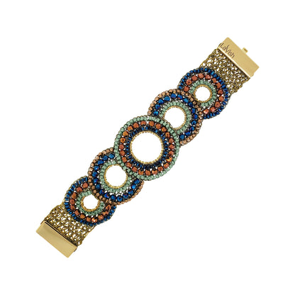 A gold-tone bracelet with a series of interlocking circular links adorned with blue, green, and orange rhinestones, shown on a plain white background. Discover the Riviera Mix Gush Bracelet, hand-crocheted in Brazil with 18K gold-filled wire, colorful crystals and an elegant circular design.
