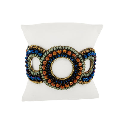 A wide beaded cuff bracelet displayed on a white pillow. The bracelet features three interlocking oval sections with intricate beadwork in blue, orange, olive green, and gold. Riviera Mix Gush handmade crochet bracelet featuring layered circles with blue, sage green and bronze crystals, gold-filled wire and magnetic clasp.