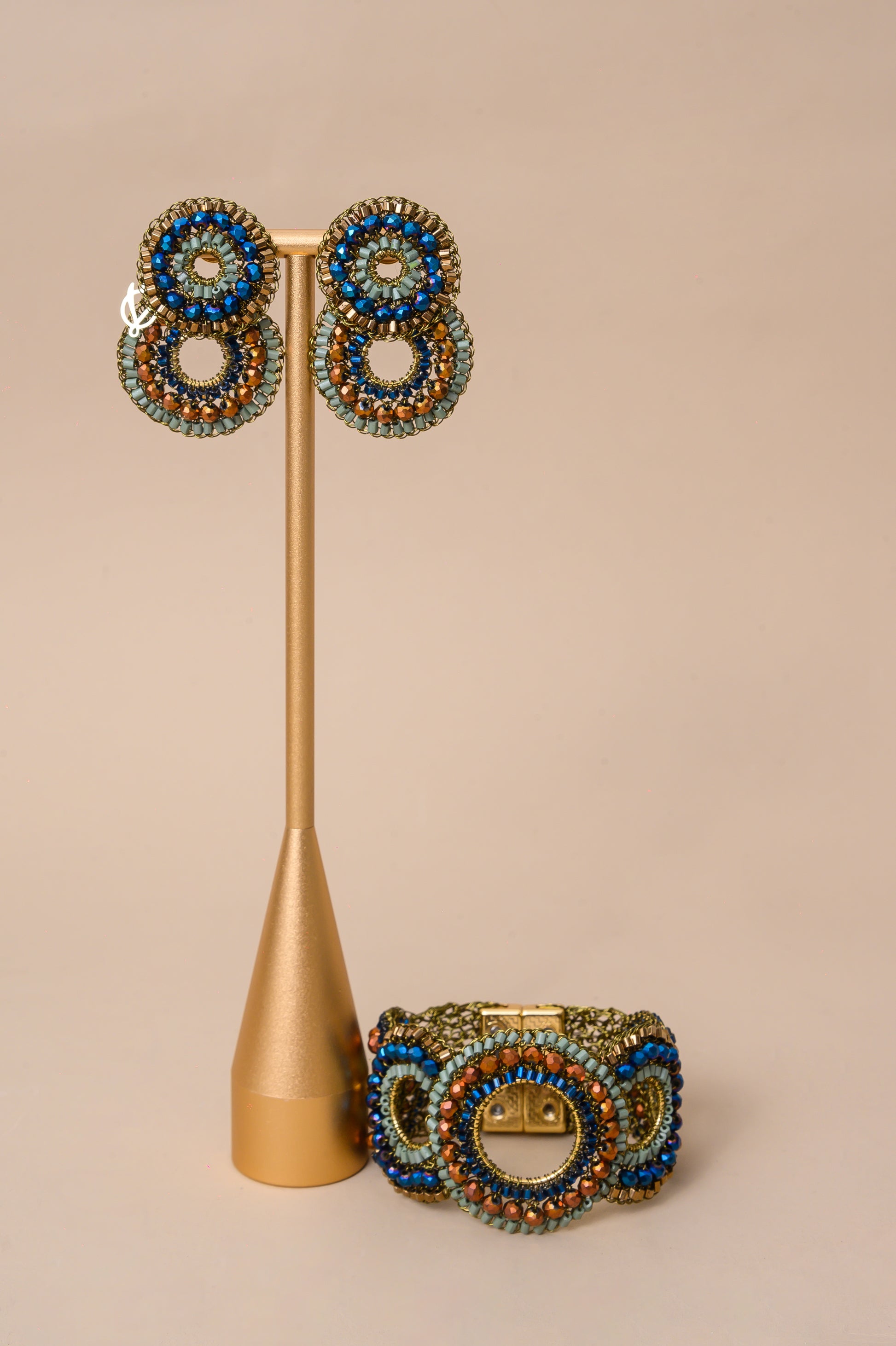A jewelry set featuring a wide beaded cuff bracelet and matching drop earrings, displayed on a gold stand against a solid beige background.