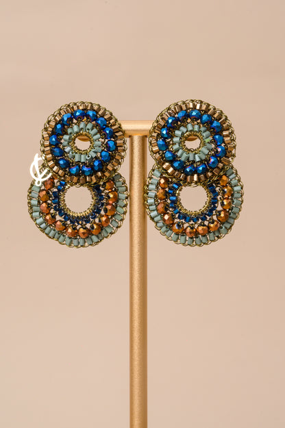 A pair of handmade beaded drop earrings displayed on a gold stand against a solid beige background. The earrings feature concentric circular patterns with blue beads, light green beads, and bronze-brown beads in a textured gold-tone metal setting.