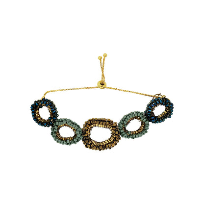 A beaded stretch bracelet featuring five interlocking oval links in shades of green, gold, and dark blue, suspended from a thin gold chain with an adjustable toggle clasp, shown on a plain white background. Shop the Riviera Mix Halo Bracelet, handcrafted in Brazil with colorful crochet links, enameled wire and an elegant adjustable design.