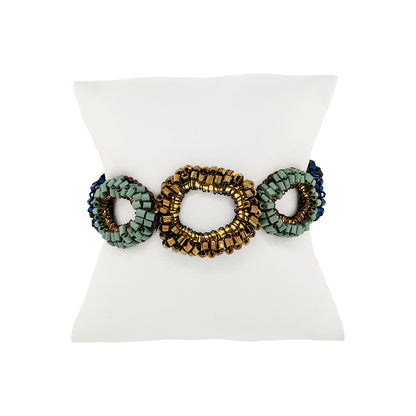 A beaded stretch bracelet with three oval links, featuring a central gold-toned link flanked by sage green and dark blue beaded oval links, displayed on a white mannequin stand.