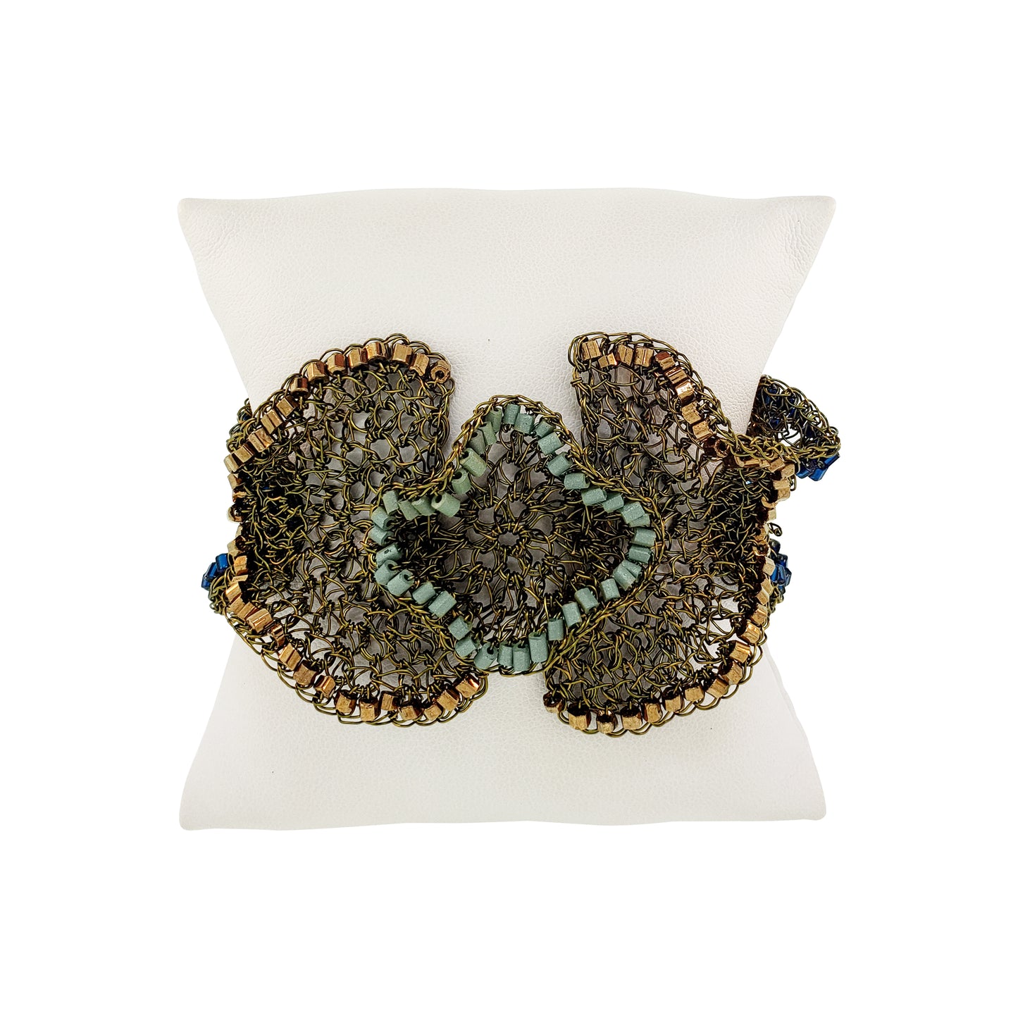 A beaded butterfly-shaped bracelet displayed on a white pillow. The bracelet features intricate metallic gold and bronze wirework with light blue-green beads and small blue bead accents. Riviera Mix Jasmine handmade crochet cuff bracelet with sage green, bronze and deep blue accents in 18 karat gold filled wire.