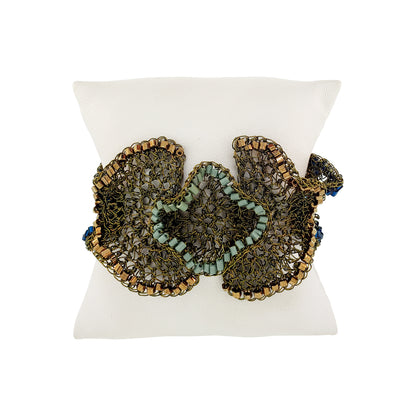 A beaded butterfly-shaped bracelet displayed on a white pillow. The bracelet features intricate metallic gold and bronze wirework with light blue-green beads and small blue bead accents. Riviera Mix Jasmine handmade crochet cuff bracelet with sage green, bronze and deep blue accents in 18 karat gold filled wire.