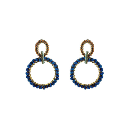 A pair of statement drop earrings featuring large open circular hoops set with deep blue faceted beads and accented with gold-toned metal and small green crystals, shown on a plain white background. Shop Riviera Mix Lola Maxi Earrings, handcrafted in Brazil with 18K gold-filled wire and sparkling blue and bronze crystals in a bold double-hoop design.