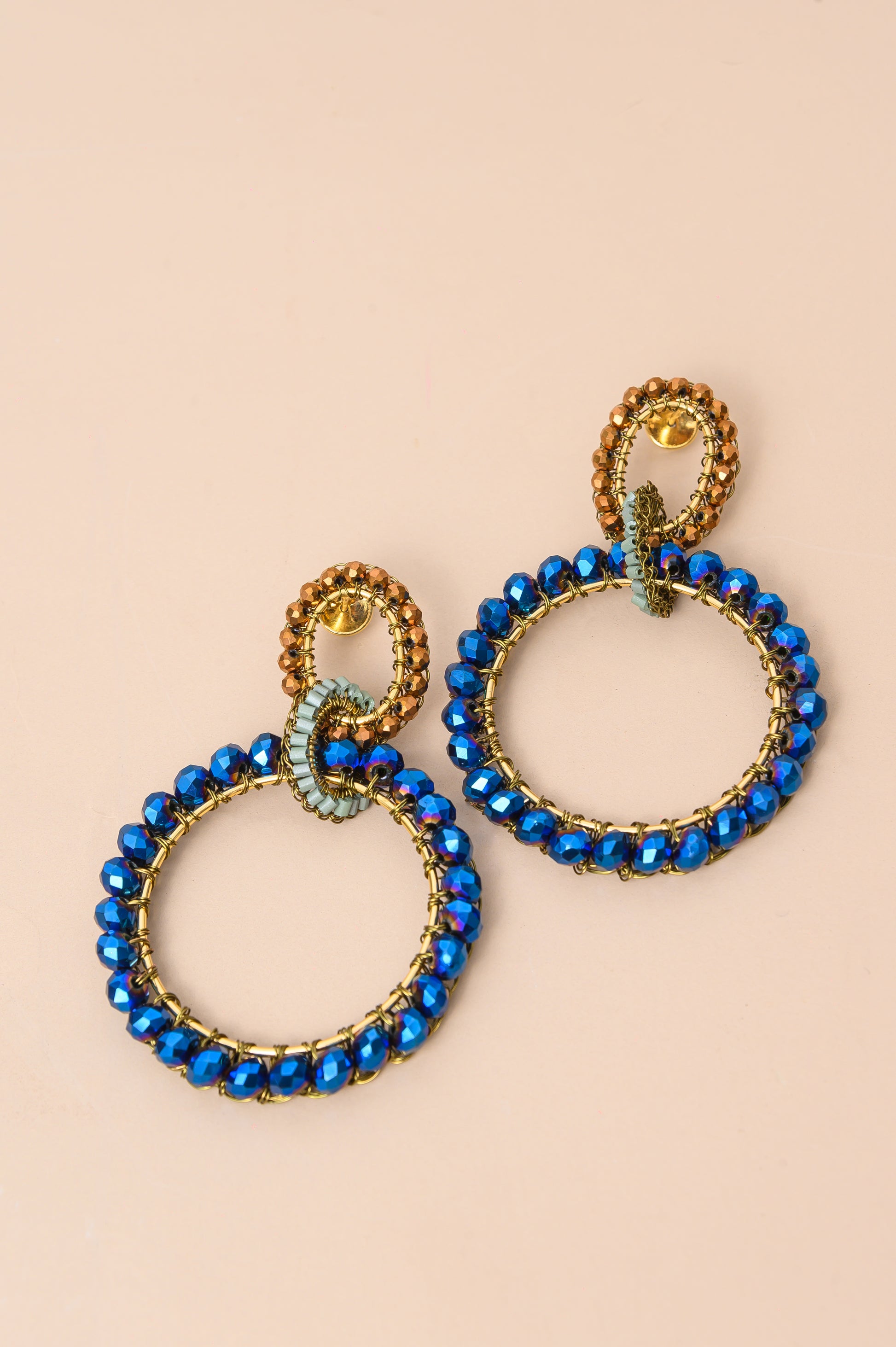 A pair of gold-tone hoop earrings with beaded blue outer rings and a central gold-and-blue link detail, displayed on a solid pale peach background. Riviera Mix Lola Maxi handmade crochet earrings with oversized blue crystal hoops, bronze accents and 18K gold-filled wire.