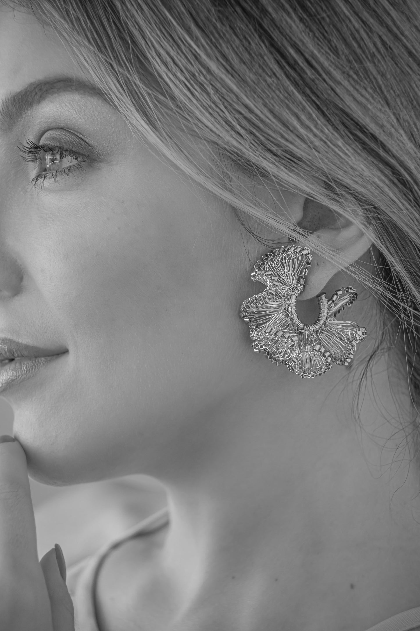 A close-up black and white photograph of a woman's profile wearing a large, textured, leaf-like statement earring. Shop Riviera Mix Rio Hoops, handcrafted in Brazil with 18K gold filled and enameled wire. Lightweight ruffled hoop earrings with colorful artisan details.