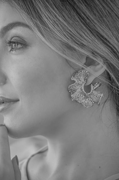A close-up black and white photograph of a woman's profile wearing a large, textured, leaf-like statement earring. Shop Riviera Mix Rio Hoops, handcrafted in Brazil with 18K gold filled and enameled wire. Lightweight ruffled hoop earrings with colorful artisan details.