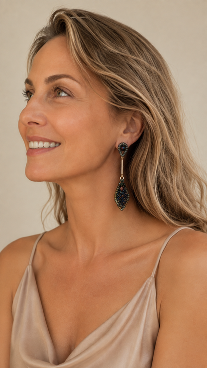 A woman with blonde hair wearing a champagne satin camisole and a long, teardrop-shaped dangle earring with dark blue and gold details, shown in a close-up profile against a warm beige background. Riviera Mix Tulip handmade crochet dangle earrings with sculptural leaf drops in deep blue, sage green and bronze with gold filled details.