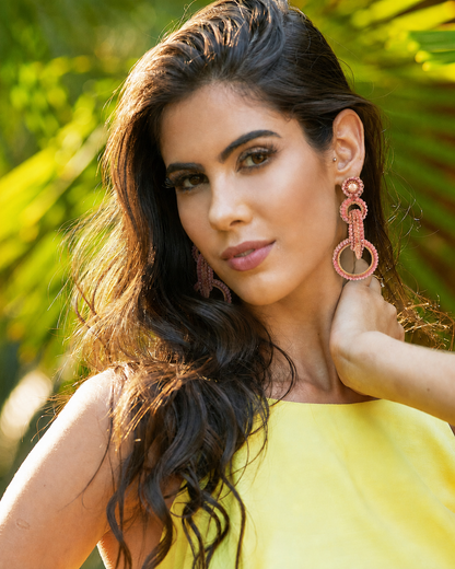 A woman with long, wavy dark brown hair wearing large, pinkish-red textured drop earrings and a bright yellow top, posing outdoors with a blurred background of green foliage. interlocked link earrings, hand-crocheted earrings, rose quartz earrings, rose gold filled earrings, 18k rose gold filled earrings, handmade Brazilian earrings, artisan statement earrings, lightweight earrings, hypoallergenic earrings, nickel free earrings, circular link earrings, wedding earrings, romantic earrings, resort earrings