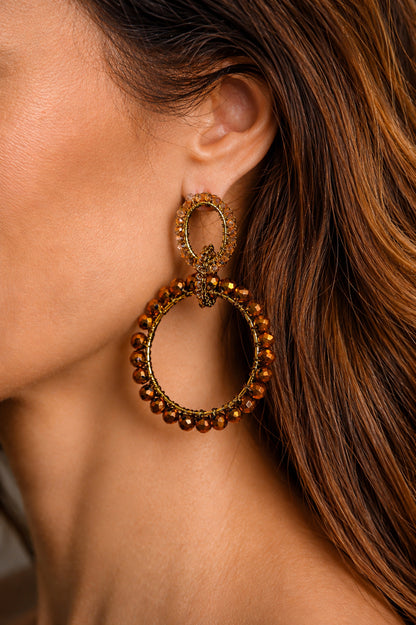 Close-up of a woman wearing a gold hoop earring with beads. Hand-crocheted double circle statement earrings with bronze crystals and 18K gold-filled and enameled copper wire, handmade in Brazil.