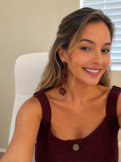 A woman wearing a pair of long, dangling burgundy earrings with textured circular details, posing in a white chair indoors.
