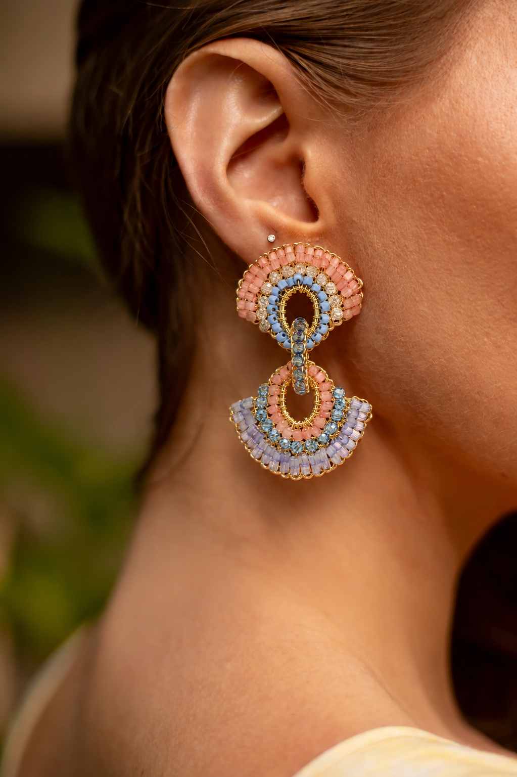 Close-up of a person wearing colorful earrings with a blurred background. Hand-crocheted in Brazil with 18K gold-filled wire, these lightweight colorful crystal earrings feature coral, blue, lavender, and gold details. Hypoallergenic, nickel-free, and perfect for statement styling.