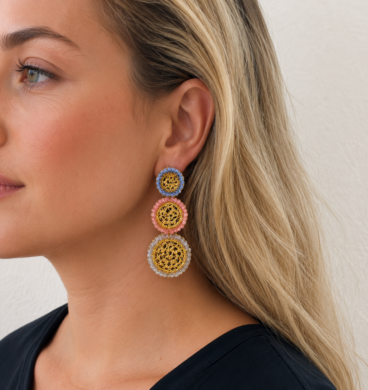 Close-up of a woman wearing colorful earrings with a neutral background. Shop handmade triple cascade mandala earrings crafted in Brazil with 18K gold-filled wire, soft blue, coral, and clear crystal accents. Lightweight, hypoallergenic, nickel-free, and perfect for special occasions or resort wear.