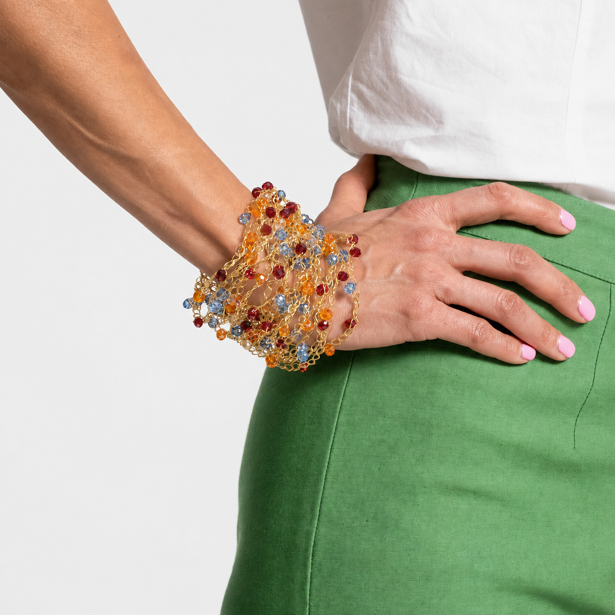 A close-up of a person's wrist and hand wearing a chunky, beaded mesh cuff bracelet in amber, red, and light blue, paired with a white top and green skirt against a light gray background. hand-crocheted cuff bracelet, gold cuff bracelet, multicolor crystal bracelet, 18k gold filled bracelet, handmade Brazilian jewelry, artisan cuff bracelet, colorful bracelet, lightweight bracelet, hypoallergenic bracelet, nickel free bracelet, magnetic clasp bracelet, resort jewelry, vacation bracelet, statement cuff brace