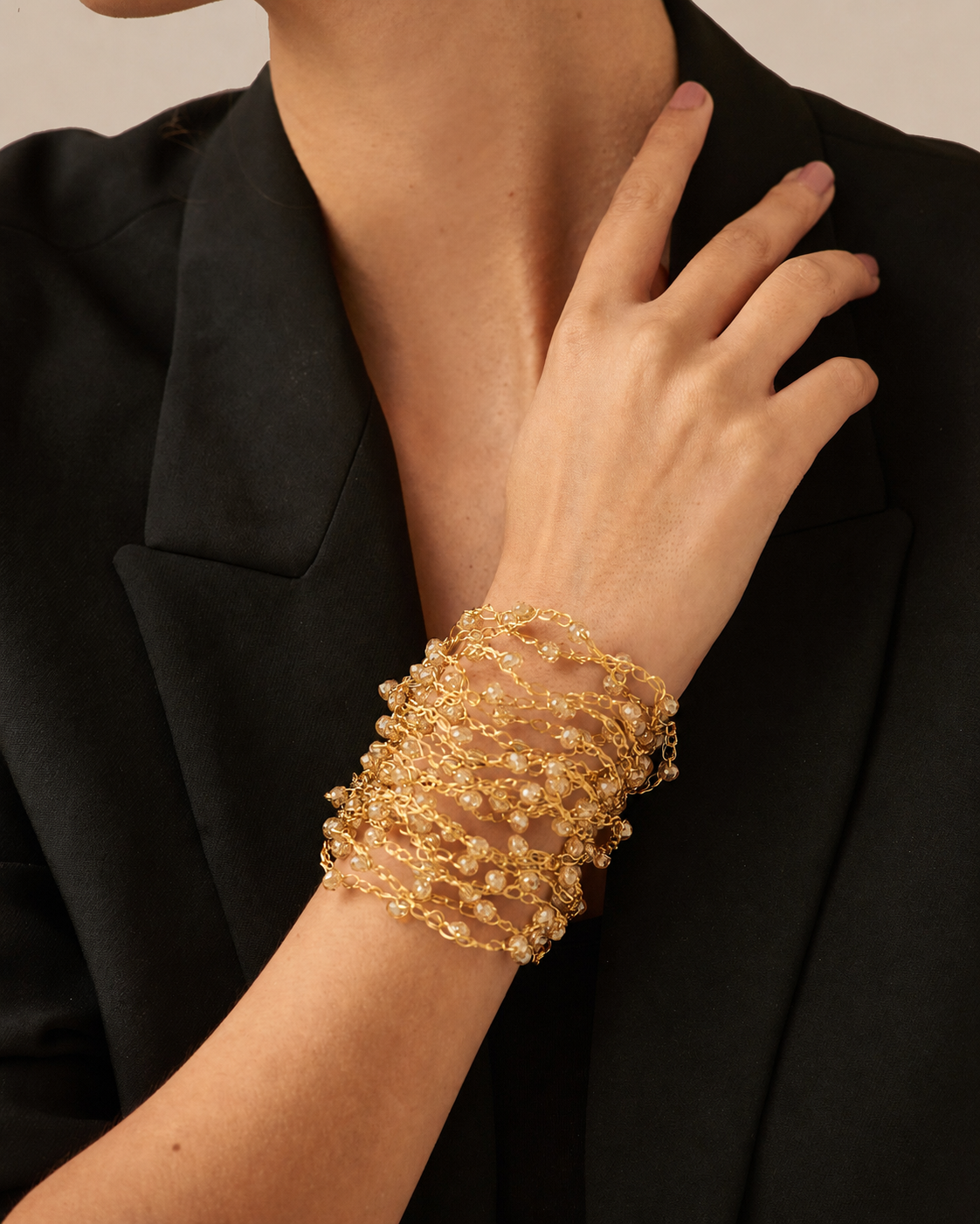 A close-up of a person wearing a wide, multi-strand cuff bracelet made of gold-toned metal and clear rhinestones, styled with a black blazer. hand-crocheted cuff bracelet, gold cuff bracelet, gold crystal bracelet, 18k gold filled bracelet, enameled wire bracelet, handmade Brazilian jewelry, artisan cuff bracelet, lightweight bracelet, hypoallergenic bracelet, nickel free bracelet, magnetic clasp bracelet, bridal bracelet, evening bracelet, resort jewelry, luxury handmade bracelet.