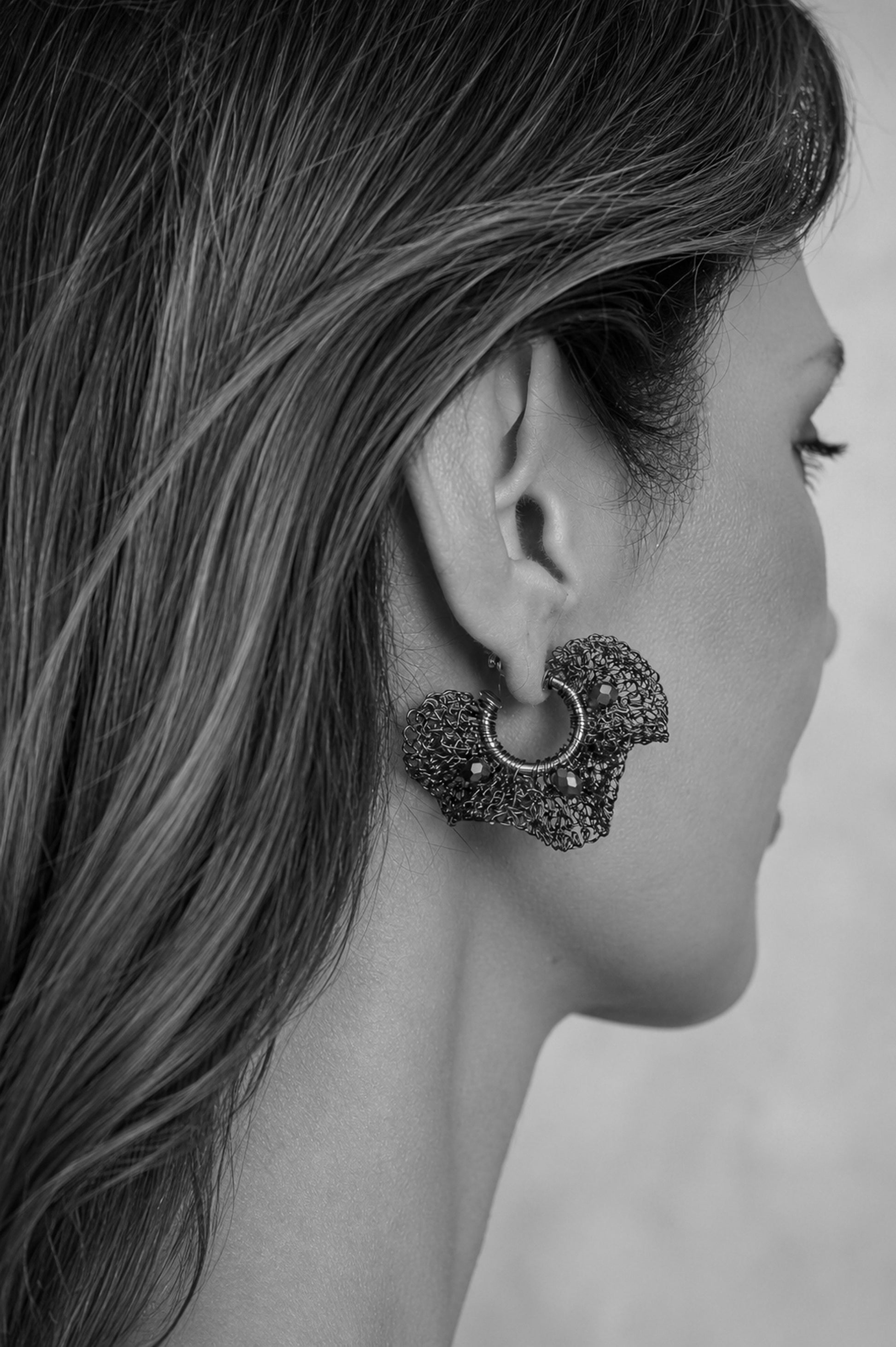 A close-up side profile of a woman wearing a textured, scalloped earring with a small hoop, shown in black and white. Nalu hand-crocheted gold hoop earrings with sculptural ruffled wirework and sparkling crystal accents, handmade in Brazil.