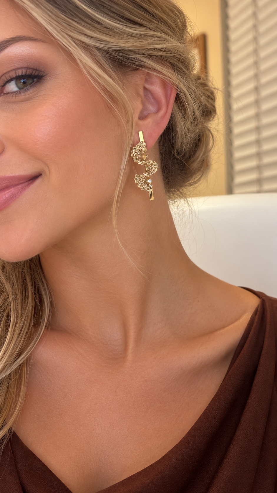 A close-up lifestyle shot of a woman wearing a gold spiral-shaped dangle earring with small clear stones, paired with a brown top. Shop handcrafted gold bar drop earrings with dimensional crochet wirework and sparkling crystals. Lightweight, hypoallergenic and handmade in Brazil.