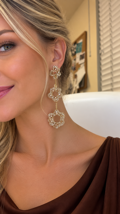 A close-up lifestyle shot of a woman wearing a pair of ornate gold drop earrings adorned with clear rhinestones. The earrings feature three stacked floral and geometric designs. Shop Ruffled Cascade Earrings, handcrafted in Brazil with 18K gold-filled and enameled wire and sparkling crystals. Lightweight, sculptural and hypoallergenic.