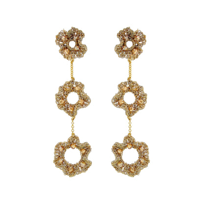 A pair of gold-tone dangle earrings with three stacked flower-shaped links, each adorned with clear rhinestones and accented with small gold beads, set against a plain white background. Hand-crocheted gold ruffled cascade earrings with three floral drops and champagne crystal accents, handmade in Brazil.