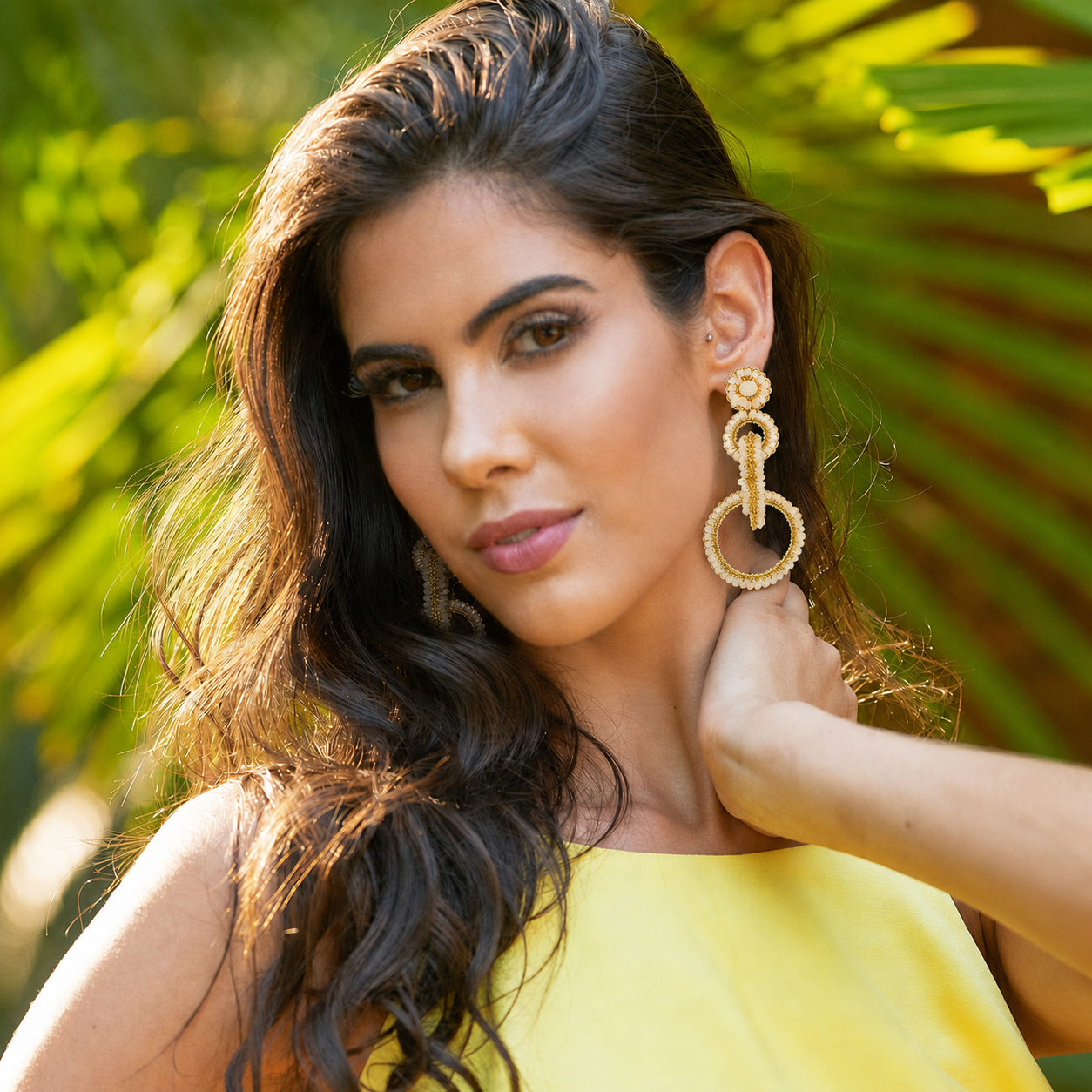 A woman with long dark wavy hair wearing large gold-toned textured hoop earrings and a bright yellow top, posed outdoors with blurred green foliage in the background. Shop interlocked link earrings handmade in Brazil with 18 karat gold filled wire, champagne-toned beads, sparkling crystals, and layered circular links. Lightweight, hypoallergenic, and nickel-free.