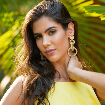 A woman with long dark wavy hair wearing large gold-toned textured hoop earrings and a bright yellow top, posed outdoors with blurred green foliage in the background. Shop interlocked link earrings handmade in Brazil with 18 karat gold filled wire, champagne-toned beads, sparkling crystals, and layered circular links. Lightweight, hypoallergenic, and nickel-free.