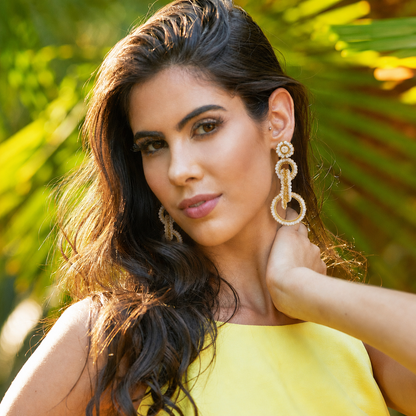 A woman with long dark wavy hair wearing large gold-toned beaded drop earrings and a bright yellow top, posed outdoors with blurred green palm leaves in the background. Shop interlocked link earrings handmade in Brazil with 18 karat gold filled wire, soft ivory beads, sparkling crystals, and layered circular links. Lightweight, hypoallergenic, and nickel-free.